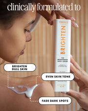 A person holds a white Truly Clinical Brighten body serum bottle while applying brightening cream to their shoulder. The image includes text overlays reading 'clinically formulated to', 'BRIGHTEN DULL SKIN', 'EVEN SKIN TONE', and 'FADE DARK SPOTS' against a warm beige background.