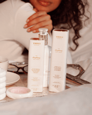 A woman sits at a table with The Face Serum Duo by Truly Beauty displayed on a tray, alongside a pair of glasses and a small pink dish.