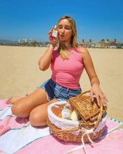 A woman in a pink top and denim shorts sits on a beach towel, making a kissy face while holding Truly Beauty’s Gabrielles Everyday Routine skincare bottle. An open picnic basket rests nearby on the sand, with buildings in the background.