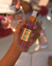 A hand with pink-painted nails holds a bottle of Truly Beauty’s Glazed Donut Perfume Mist. In the blurred background are a wine glass, flowers, and an open book, evoking the mists irresistible toasted vanilla and gourmand scent.