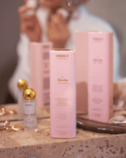 A bottle of Truly Beauty’s The Face Serum Duo rests on a bathroom counter next to its pink packaging, jewelry, and other skincare items, while someone in the background applies this brightening serum.