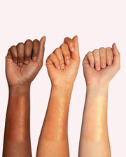 Three raised forearms of varying skin tones display swatches of Truly Beauty’s Sun Drops Bronze Body Glow, showcasing its hydrating, shimmery golden finish and sunkissed radiance against a pale background.
