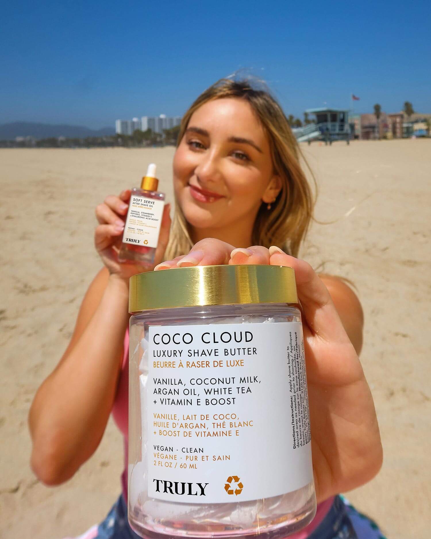 At the beach, a woman holds Gabrielles Ultimate Routine by Truly Beauty toward the camera with its hydrating formula visible. In her other hand is a dropper bottle, and buildings and sand appear in the background.