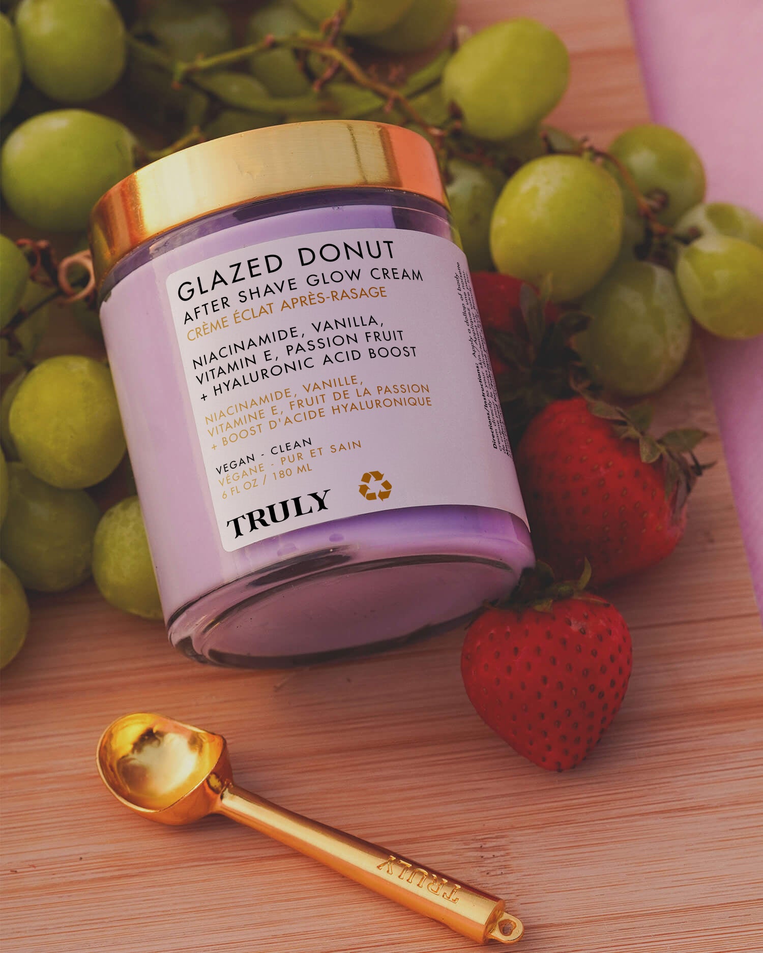 A jar of Truly Beauty’s Glazed Donut Shave Duo rests on a pink surface with green grapes and strawberries nearby, offering razor burn relief. A small gold spoon is placed in front of the jar.