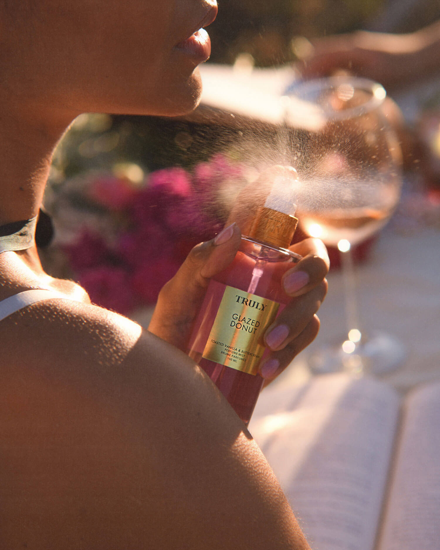 A person sprays Truly Beauty Glazed Donut Perfume Mist onto their neck as sunlight illuminates the mist. A wine glass and blurred flowers in the background evoke the gourmand scent of toasted vanilla and whipped buttercream.