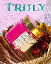 A pink jar labeled “Kaylas Everyday Routine” by Truly Beauty sits in a golden dish beside fresh cherries, with a towel embroidered “TRULY” in the background—perfect to pair with Soft Serve Whipped Shave Butter for silky smooth skin.