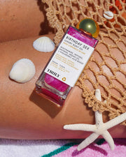 A bottle of Zaras Everyday Routine by Truly Beauty sits on a persons leg, wrapped in crochet, with Buns of Glowry Butt Butter nearby and seashells and a starfish scattered on sun-kissed skin.