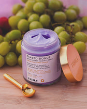 A jar of Truly Beauty’s Glazed Donut Shave Duo with light purple cream sits on a wooden surface. A gold lid and spoon are nearby for application and razor burn relief, while green grapes add a fresh touch to the scene.