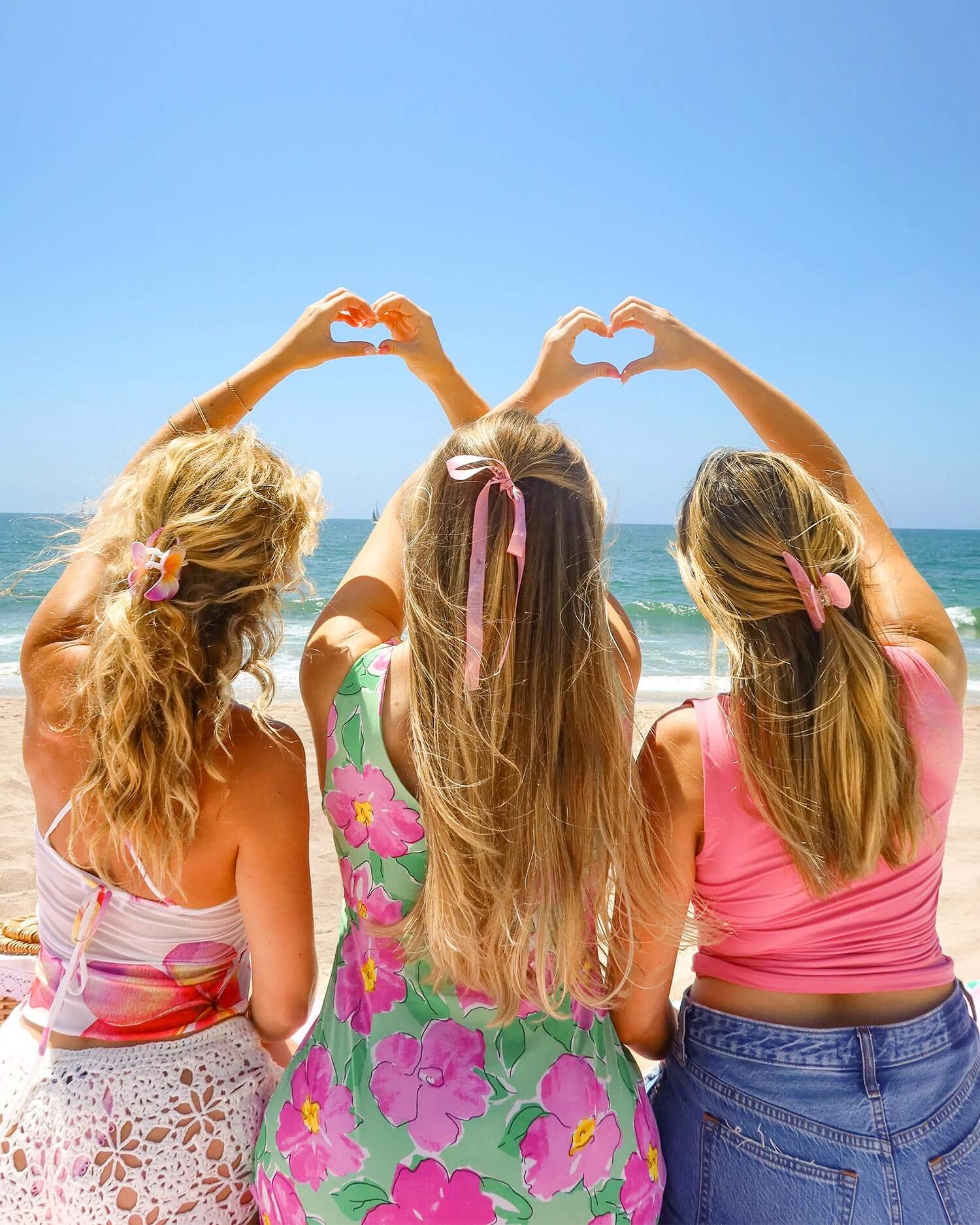 Three blonde women in summer outfits and pink ribbons sit on the beach, their skin glowing from Zaras Everyday Routine by Truly Beauty. Two make heart shapes with their hands against the blue sky as they enjoy the ocean view.