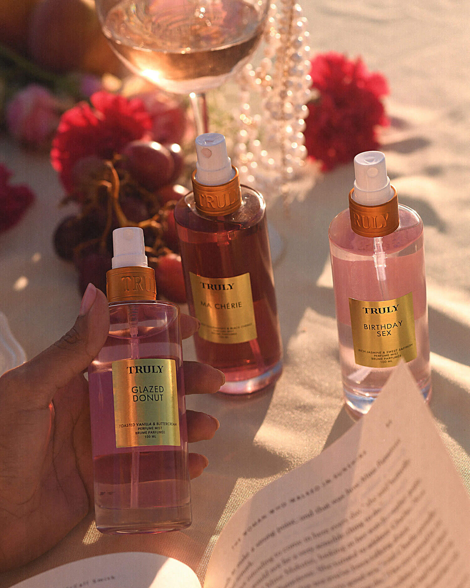 A hand holds a bottle labeled Truly Glazed Donut spray. Nearby, two more Truly Beauty sprays—Unicorn Chérie and Birthday Sex Perfume Mist—rest on a sunlit table amidst flowers, grapes, wine, and an open book.
