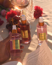 A hand holds a bottle labeled Truly Glazed Donut spray. Nearby, two more Truly Beauty sprays—Unicorn Chérie and Birthday Sex Perfume Mist—rest on a sunlit table amidst flowers, grapes, wine, and an open book.