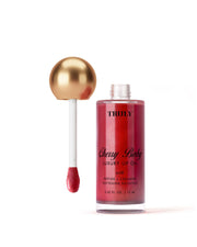 A bottle of Truly Beauty Luxury Peptide Lip Oil with a gold-topped applicator wand stands upright. The shimmering red lip oil is set against a soft white background.