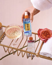 A hand holds a blue bottle of Clean Girl Perfume Mist by Truly Beauty beside a glass of pale liquid, a Glazed Donut skin care product, a pink rose, and a white towel on a gold wire bath tray—suggesting clean fragrance and fresh florals.