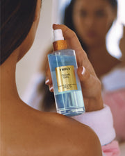 A person holds a Truly Beauty Clean Girl Perfume Mist with a gold cap. The softly blurred background suggests fresh florals and clean fragrance, while their shoulder and mirror reflection are visible.