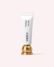 A white tube of Truly Beauty’s Hydrating Lip Gloss with black text stands upright on a round, gold metallic base against a light pink background, highlighting its high-shine formula with hyaluronic acid.