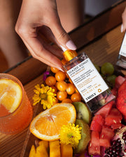 A hand holds a bottle of Truly Beauty’s Glazed Donut After Shave Oil above a tray of sliced orange, kiwi, and mango—ideal for hydrating skin and preventing ingrown hairs after your daily shave.