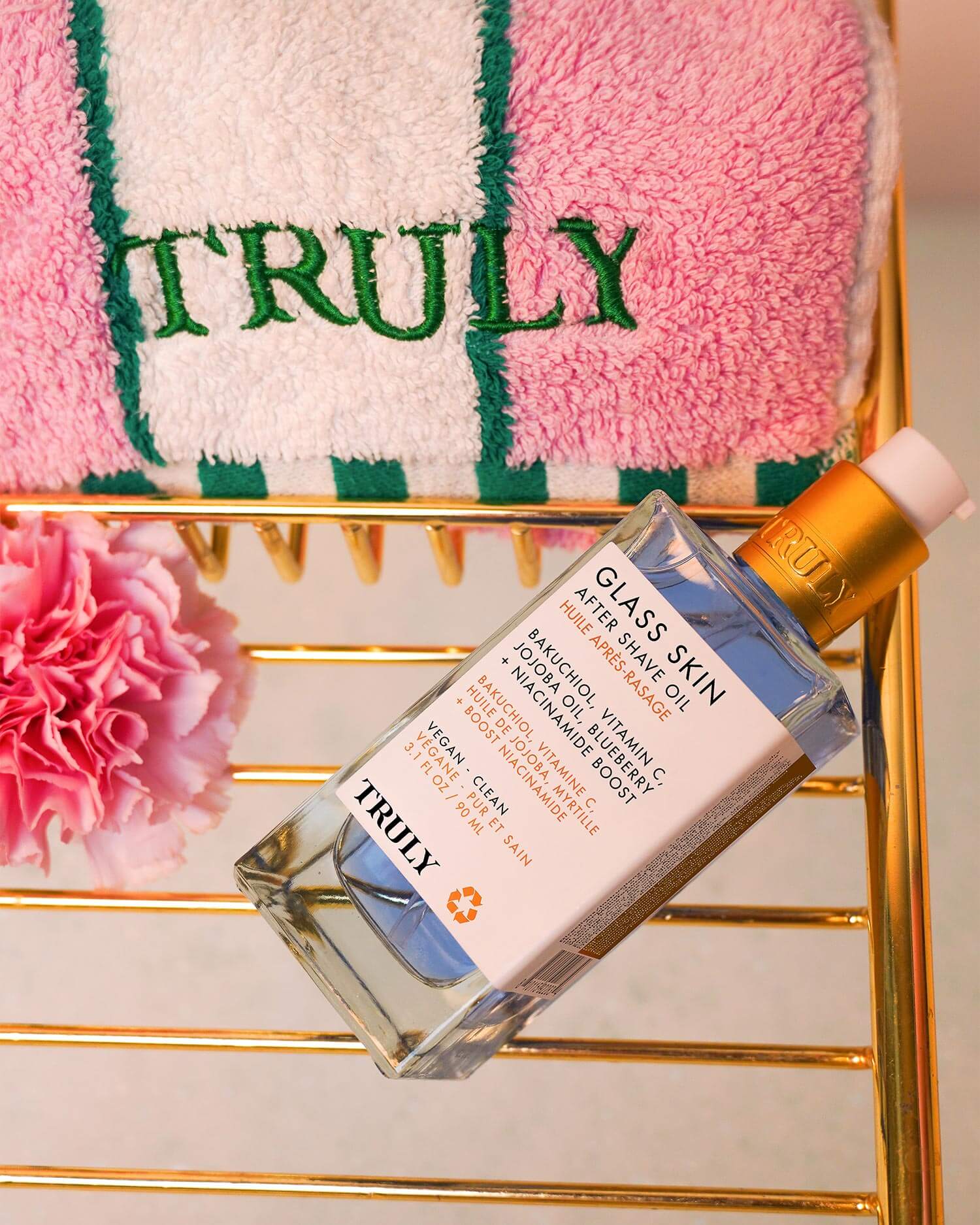 A bottle of Truly Beauty Glass Skin After Shave Oil with a gold cap sits on a gold rack beside a pink carnation and a folded pink and white towel embroidered with TRULY.