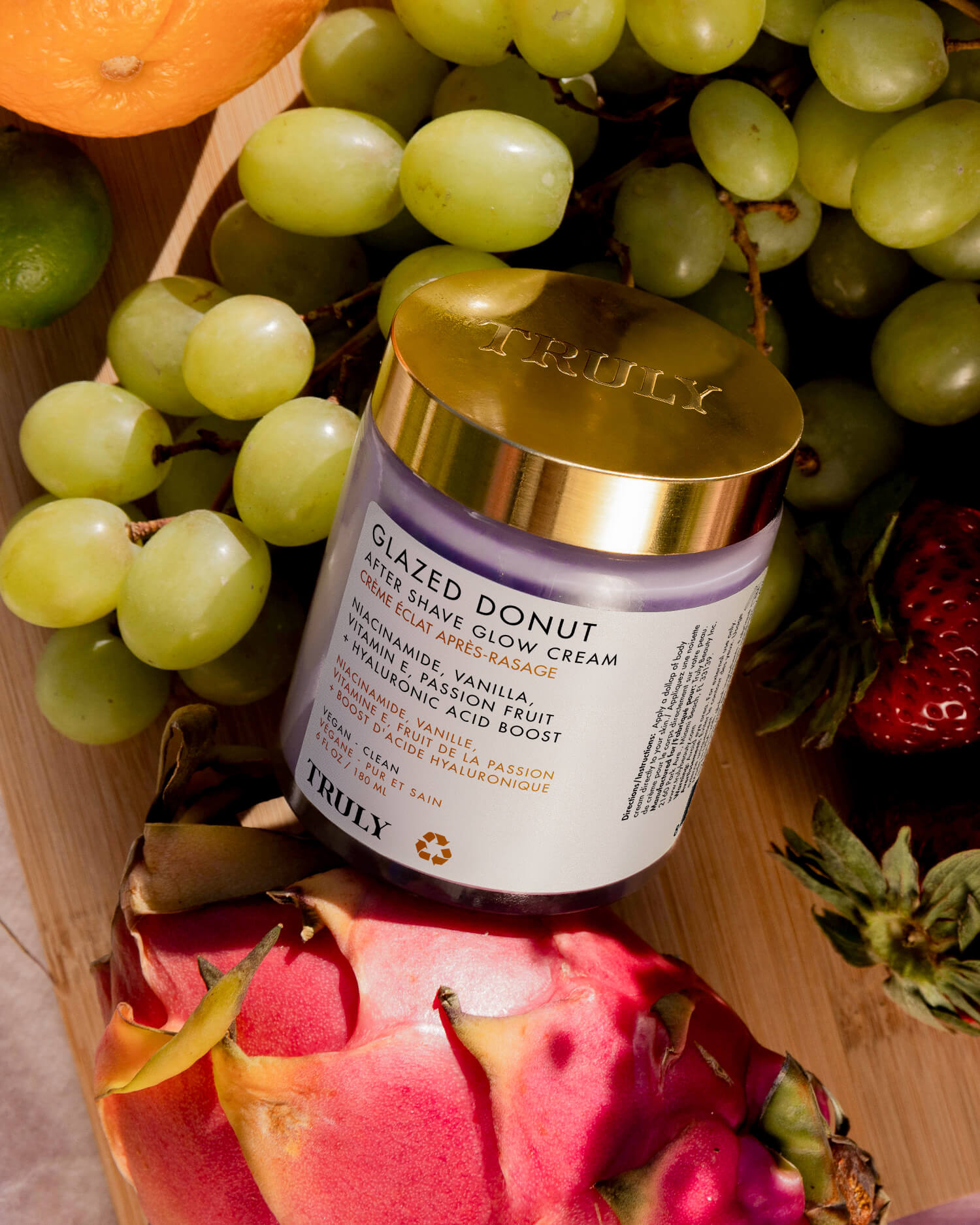 A jar of Truly Beautys Glazed Donut After Shave Glow Cream with a gold lid, surrounded by green grapes, dragon fruit, and a strawberry, creates an irresistible and vibrant appeal for this body cream.
