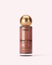 A bottle labeled Truly Beauty The Glossy Duo, featuring a gold spherical cap and filled with pinkish-brown liquid, is displayed against a pale pink background.
