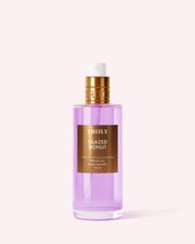 A bottle of Truly Beauty’s Glazed Donut Perfume Mist with a gold cap and pink liquid stands upright on a light pink background, emitting toasted vanilla notes for a dreamy gourmand fragrance experience.
