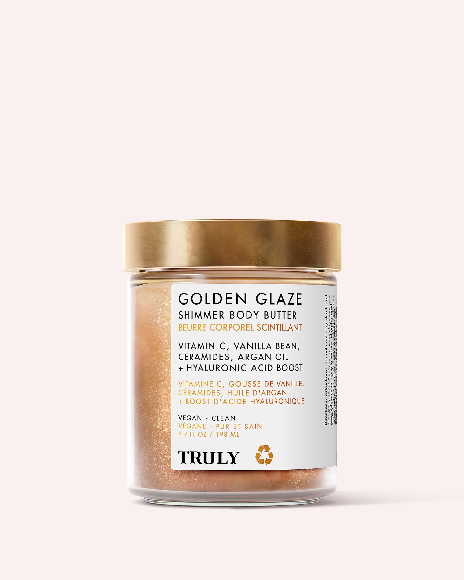A clear jar with a gold lid holds Truly Beauty’s Golden Glaze Shimmer Body Butter, featuring labels that highlight hydrating and brightening ingredients like vitamin C, vanilla bean, ceramides, argan oil, and hyaluronic acid.
