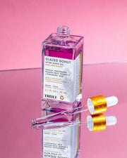 A clear bottle of Truly Beauty Glazed Donut After Shave Oil filled with purple liquid sits on a reflective surface, flanked by two glass droppers with gold caps. The pink background highlights this hydrating daily shave serum.