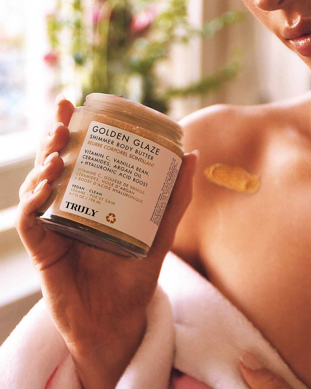 A person applies golden shimmer body butter from the Golden Glaze Luxury Trio by Truly Beauty to their shoulder, wrapped in a soft pink and white towel.