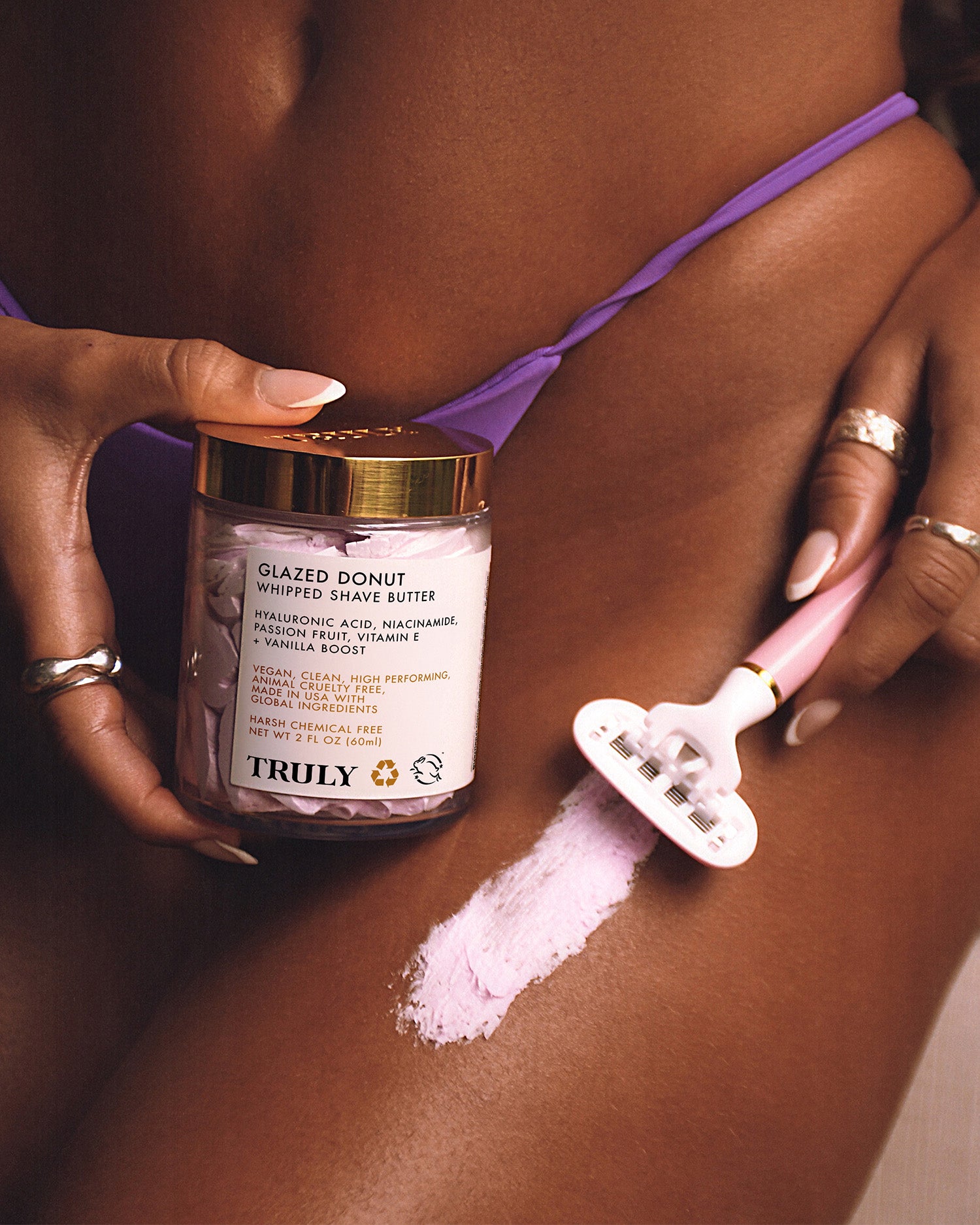 Wearing silver rings and a purple bikini, a person applies Truly Beauty’s Glazed Donut Shave Duo to their upper thigh with a razor, revealing smooth skin and razor burn relief. The product jar and label are clearly visible.