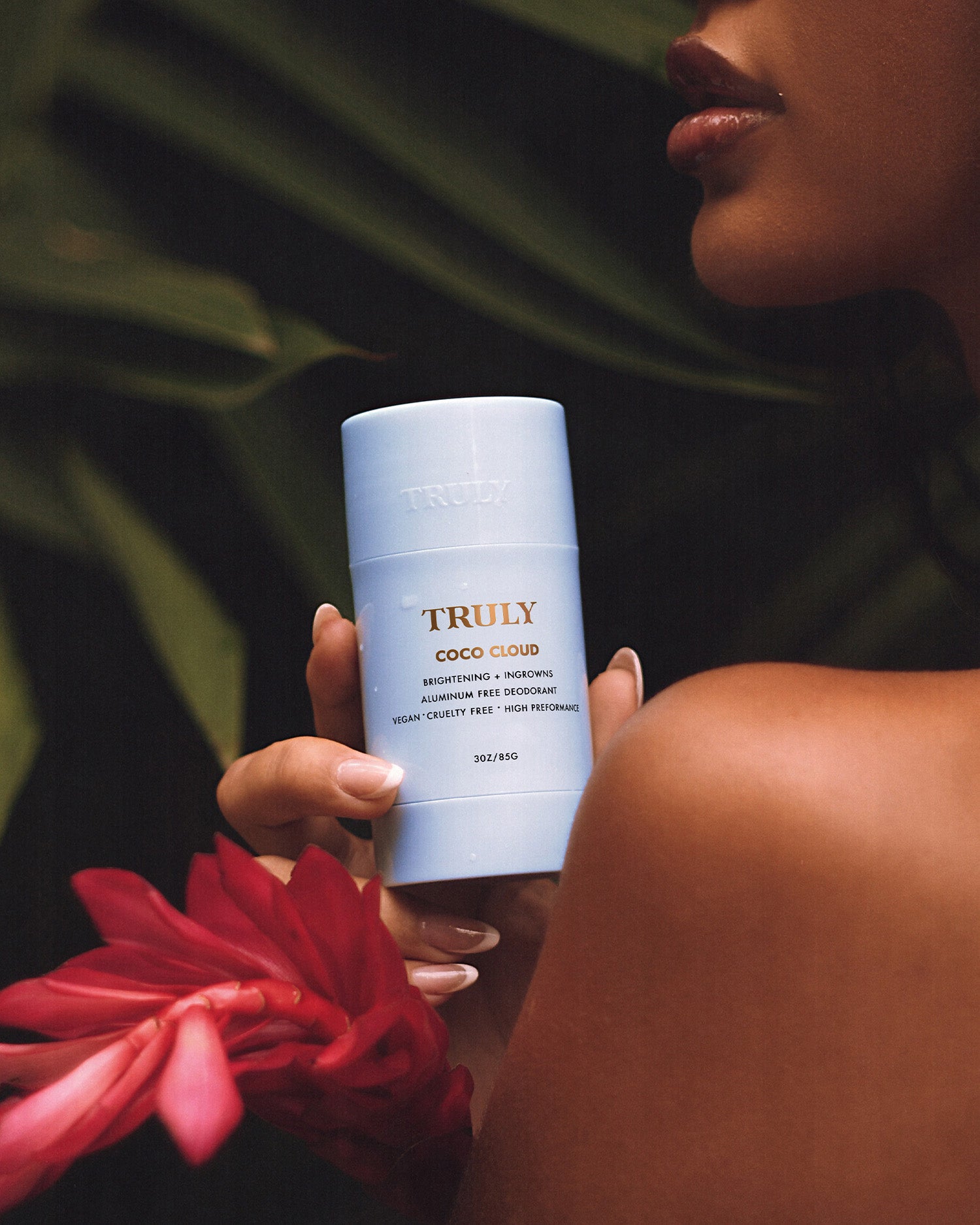 A hand holds a light blue Coco Cloud Ingrown Prevention and Brightening Deodorant by Truly Beauty with a red tropical flower. Green leaves form the background, and part of the person’s face is visible.