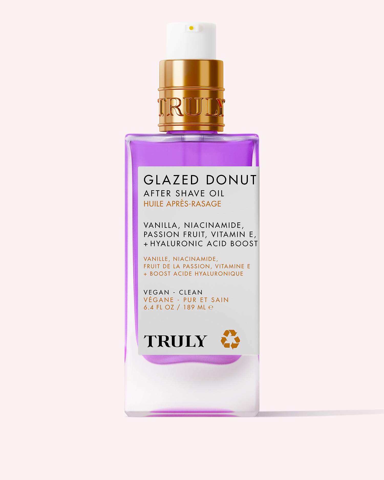 A rectangular bottle of Glazed Donut After Shave Oil with a gold pump and white label stands against a pale pink background. The light purple oil inside helps smooth and soften skin after shaving.