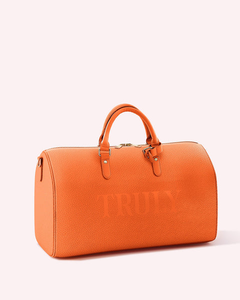 The Truly Beauty Truly Fall Duffle Bag is an orange leather travel bag with two handles, a top zipper, and TRULY embossed on the side—ideal for fall style, shown against a light pink background.