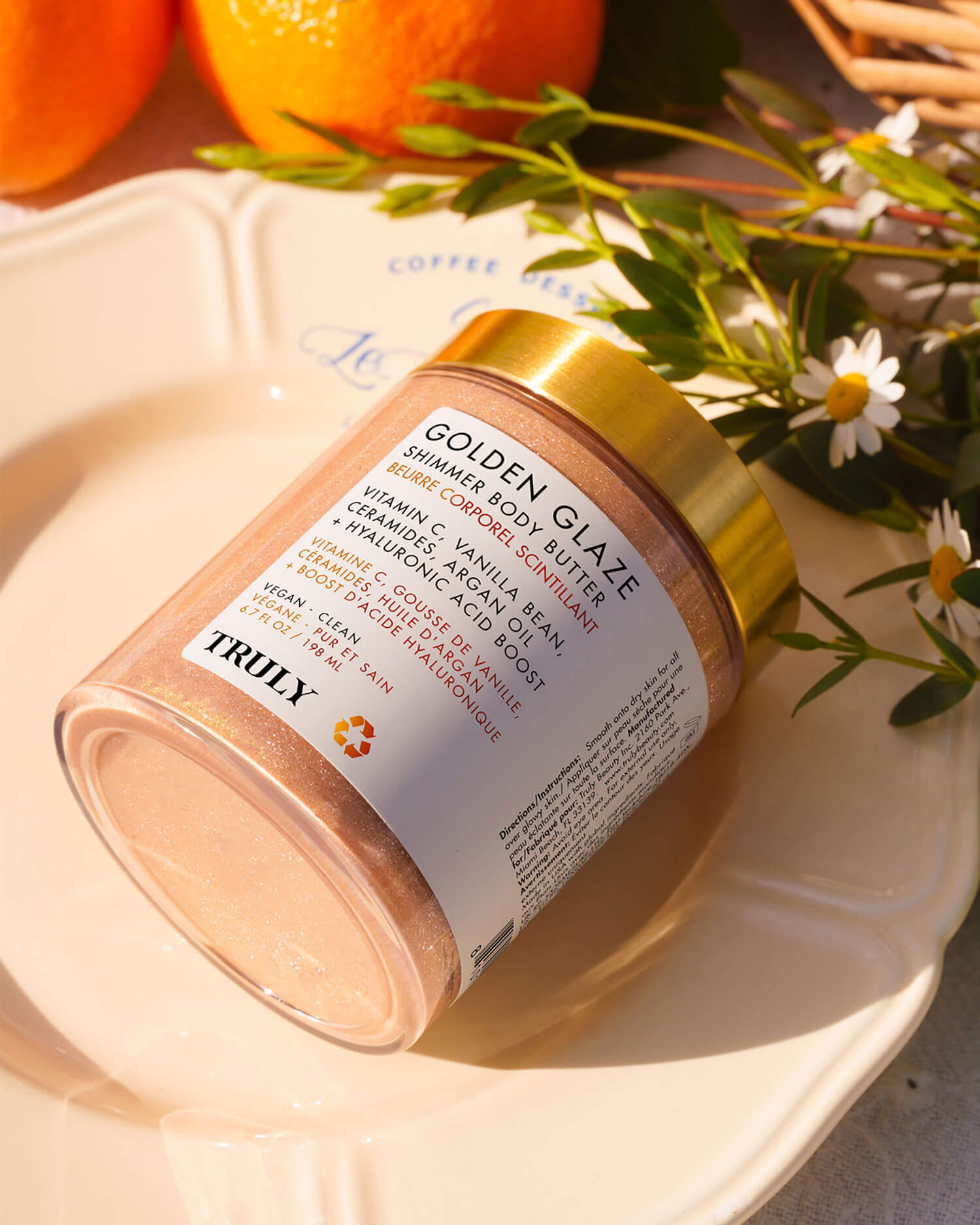 A jar of Truly Beauty Golden Glaze Shimmer Body Butter with a gold lid sits on a cream plate, surrounded by oranges and daisies in warm sunlight.