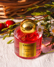 A pink glass bottle of Truly Beautys Cherry Baby Eau De Parfum with a shiny gold cap rests on a white textured surface, surrounded by jasmine flowers and greenery, with a wicker basket in the background.