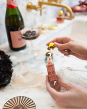 A person’s hands hold a dropper and a small bottle from Truly Beauty’s The Glossy Duo over a marble counter with hydrating lip oil, a glass of dark beverage, a champagne bottle, and gold fixtures in the background.