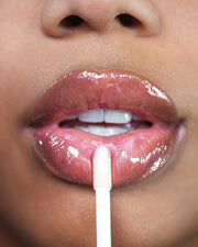 A close-up of someone applying Truly Beauty’s The Glossy Duo lip gloss with a sponge applicator shows their slightly parted lips and teeth, highlighting the shiny, plumping finish.