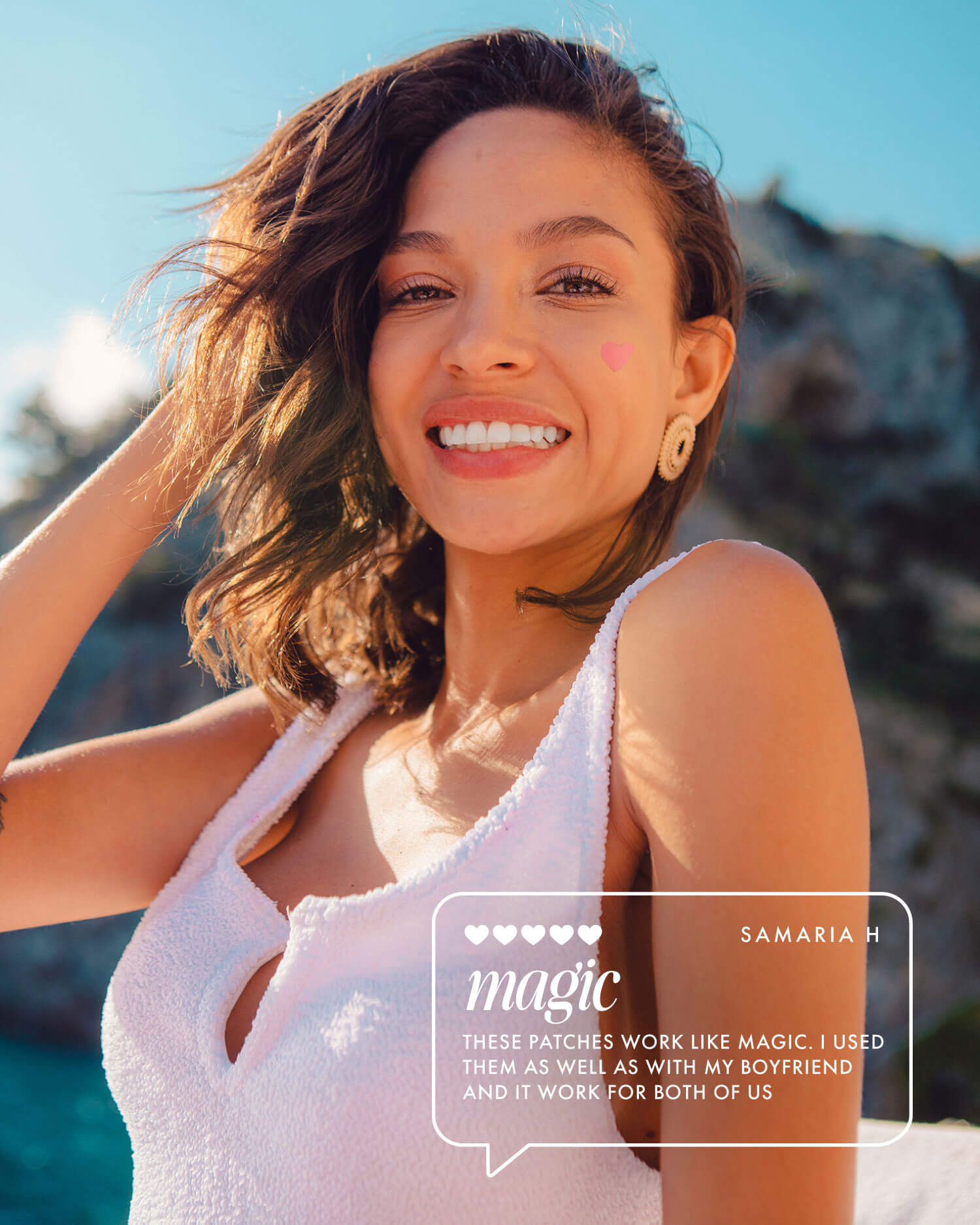 A smiling woman with short brown hair, in a white sleeveless top and heart-shaped earrings, stands outdoors with a pink heart sticker on her cheek. She shares a testimonial about her Truly Beauty Smooth Legend - Complete Collection shaving routine.
