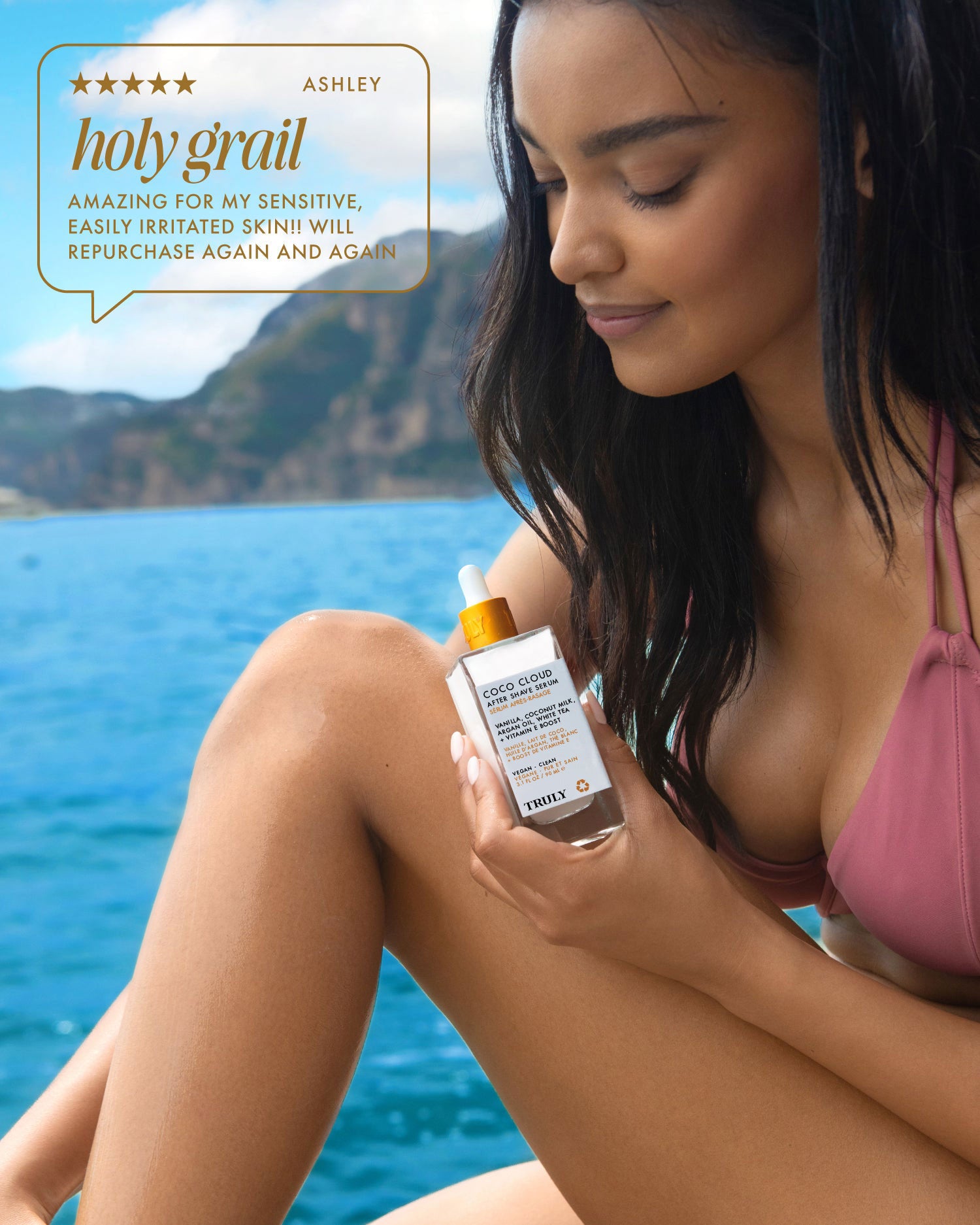 A woman in a mauve bikini relaxes by the water, holding a bottle of Truly Beauty’s Coco Cloud After Shave Serum. A review bubble says: holy grail—amazing for my sensitive, easily irritated skin! Will repurchase again and again.