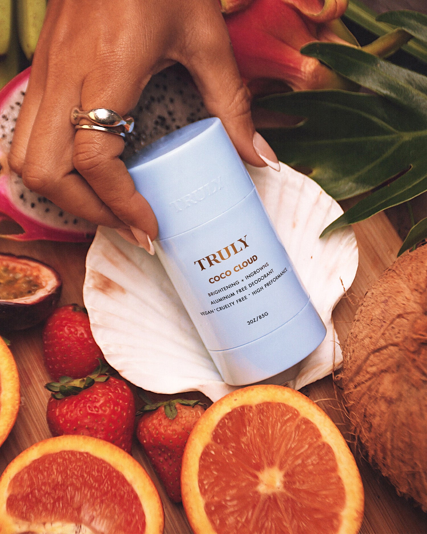 A hand holds the Coco Cloud Ingrown Prevention and Brightening Deodorant by Truly Beauty, resting on a seashell among tropical fruits, promising brighter underarms with this aluminum-free formula.