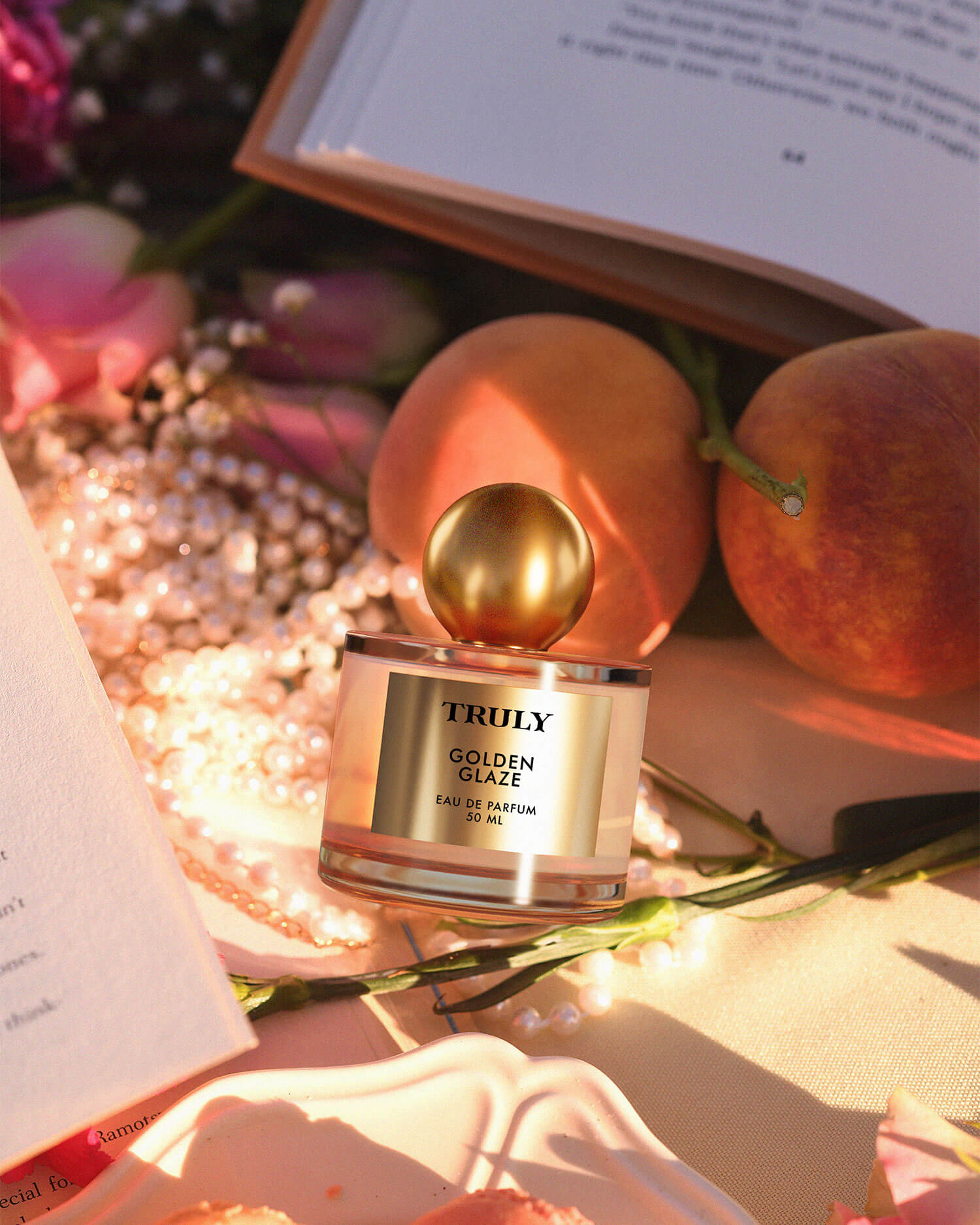 The Golden Glaze Luxury Trio by Truly Beauty rests on a beige surface with pink roses, peaches, pearls, and open books in warm sunlight—offering a hydrating scent that radiates youthful elegance.