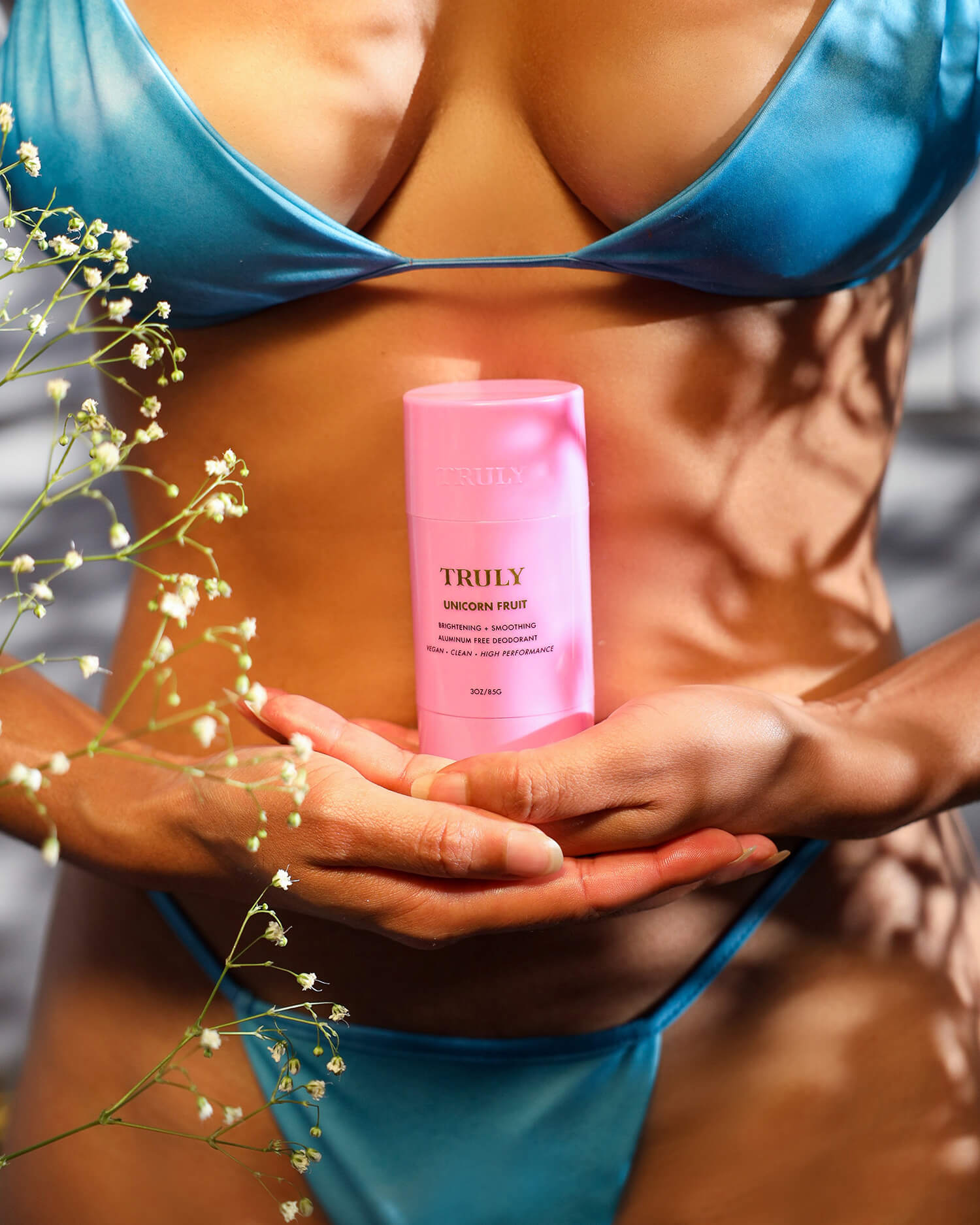 A person in a shiny blue bikini holds a pink Truly Beauty Unicorn Fruit Skin Brightening and Smoothing Deodorant, with white babys breath flowers in the foreground, showcasing this aluminum-free underarm care essential.