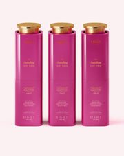 Three pink, cylindrical bottles of Truly Beauty The Smoothing Body Serum (3.1 fl oz/90 ml each) with gold caps are lined up side by side on a pale pink background. This serum targets rough, bumpy skin and keratosis pilaris.