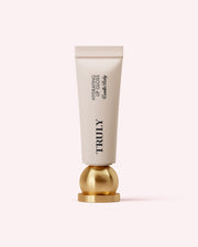 A white tube of Truly Beauty Hydrating Lip Gloss with black text stands upright on a shiny gold sphere against a pale pink background, highlighting its high-shine finish and hydrating hyaluronic acid formula.