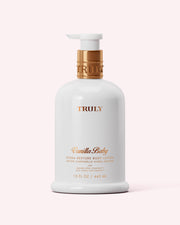 A white 15 fl oz (443 ml) bottle of Truly Beauty Hydra-Restore Body Lotion with gold pump and lettering, featuring an Amino-Lipid Complex for lasting hydration, shown against a pale pink background.