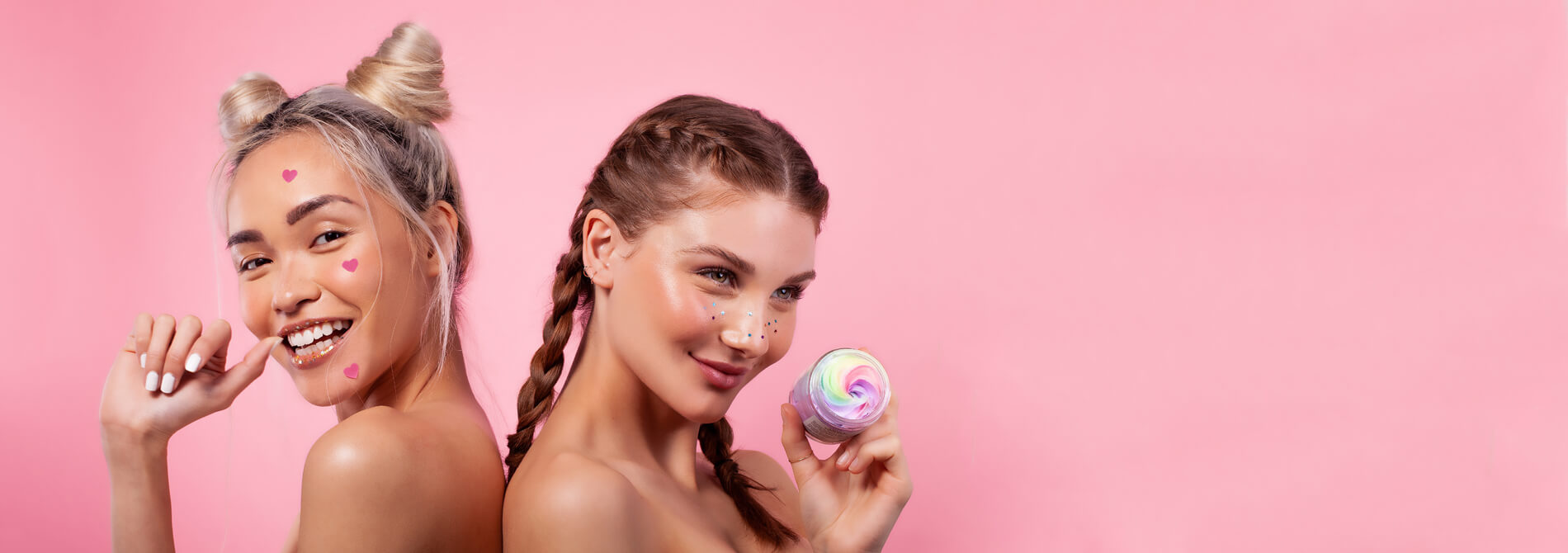 Two young women with playful hairstyles and colorful face paint pose against a pink backdrop for the Truly About Us banner; one holds a jar of Truly’s cream, both smiling brightly at the camera.