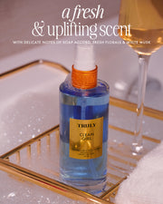 A blue bottle of Clean Girl Perfume Mist by Truly Beauty sits on a gold bath tray with bubbles, a champagne glass, and a white towel. The text highlights its clean scent with fresh florals, soapy notes, and white musk.