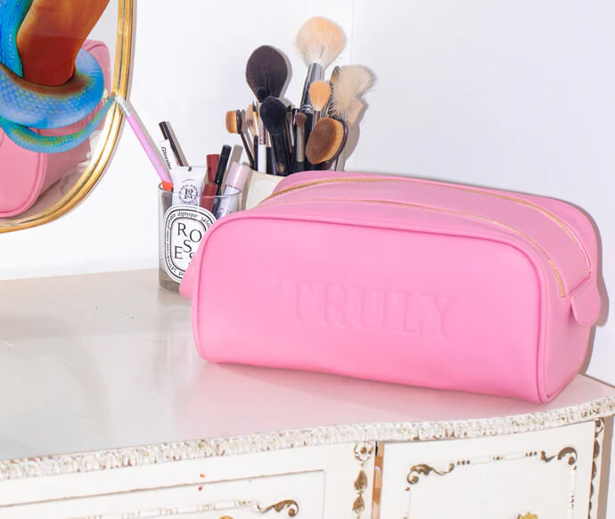 A pink makeup bag with TRULY embossed on it sits on a white vanity next to a glass holding makeup brushes and pens, with part of a mirror and a colorful object visible on the left.