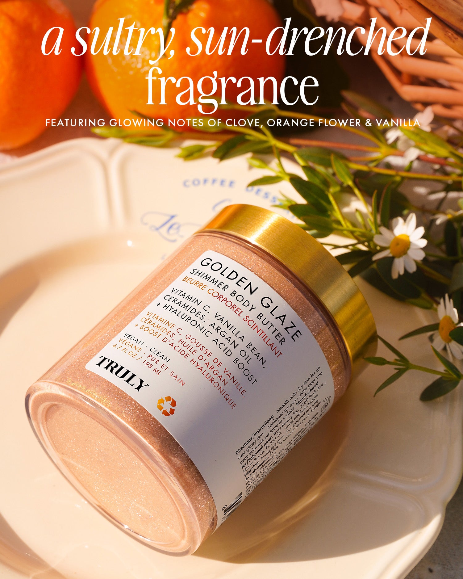 A jar of Truly Beautys Golden Glaze Shimmer Body Butter sits on a plate with flowers, orange slices, and sunlight, featuring text about its hydrating clove, orange flower, and vanilla fragrance.