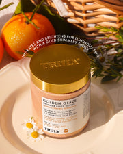 A jar from the Golden Glaze Luxury Trio by Truly Beauty sits on a white plate with oranges and a wicker basket behind, its label promoting hydration and brightening for luminous, shimmery skin.