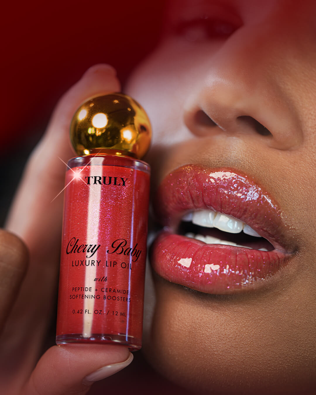 Red lip oil with gold cap held close to a person's lips against a red background