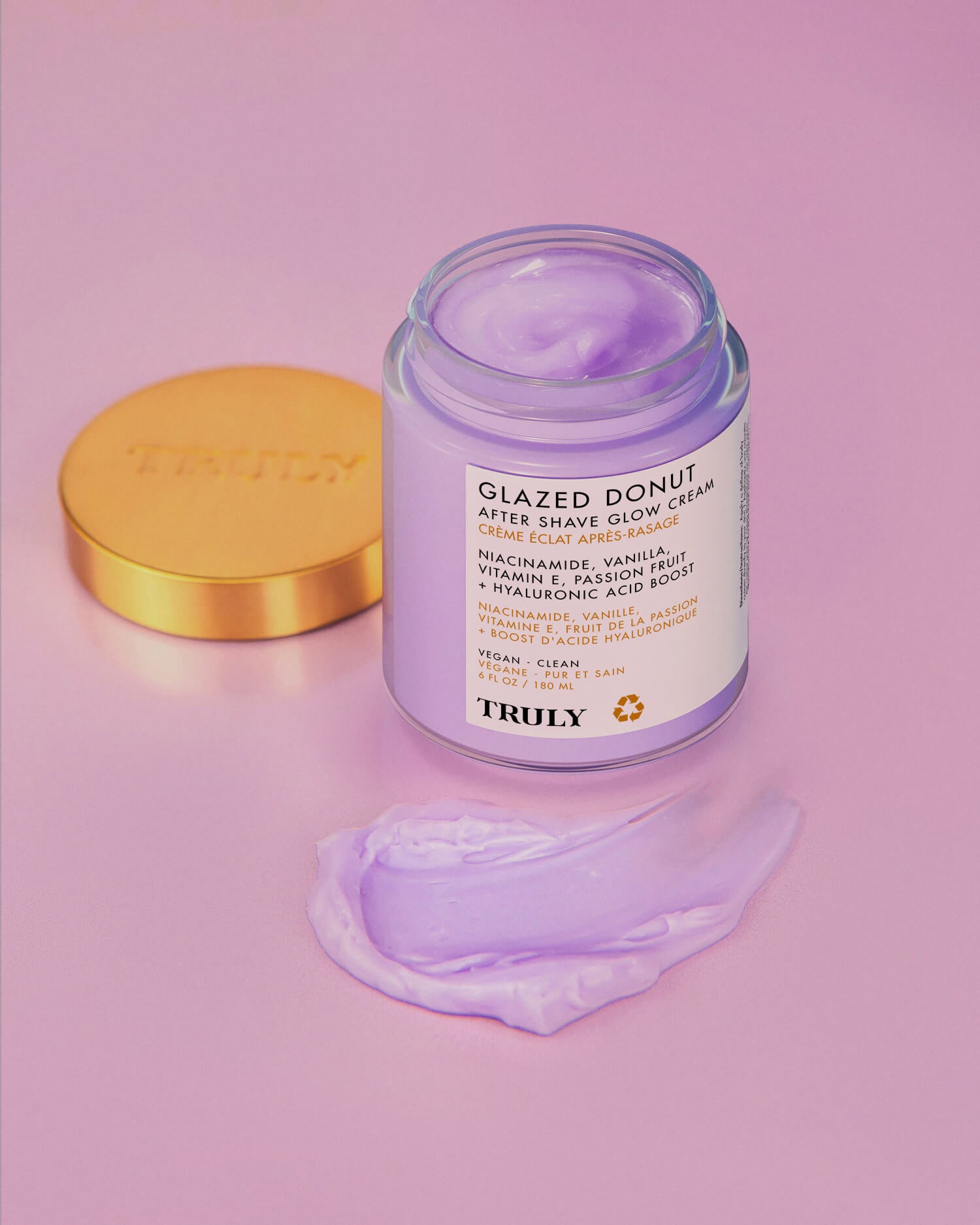 A jar of Truly Beauty’s Glazed Donut After Shave Glow Cream with an open gold lid rests on a pink surface, with some light purple cream smoothly spread in front of it.